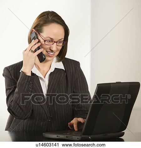 Stock Image - Caucasian businesswoman sitting at desk smiling with laptop computer talking on cellphone.. Fotosearch