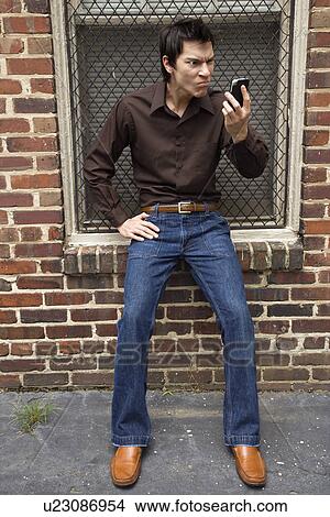 Confused young Asian man next to brick wall and window looking at cell phone. View Large Photo Image Picture - Confused young Asian man next to brick wall and window looking at cell phone.. Fotosearch