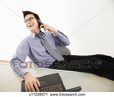 Stock Image - Laughing Asian businessman sitting at desk talking on cellphone.. Fotosearch