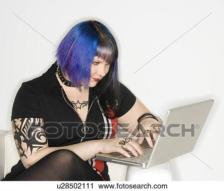 Stock Image - Portrait of Caucasian woman with blue hair, tattoo, and spike collar typing on laptop against white background.. Fotosearch