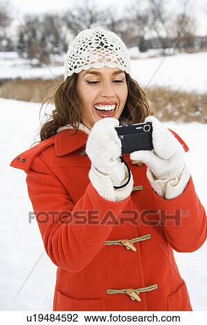 Stock Image - Smiling young adult female in winter clothing using digital camera outdoors. Fotosearch