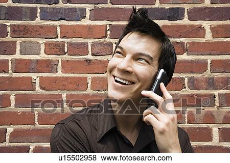 Stock Photo - Smiling young Asian man next to brick wall talking on cell phone.. Fotosearch