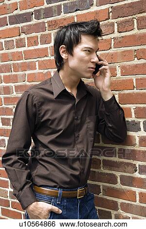 Stock Photograph - Young Asian man next to brick wall talking on cell phone.. Fotosearch