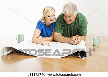 Stock Image - Middle-aged couple lying on floor looking at and discussing architectural blueprints together.. Fotosearch