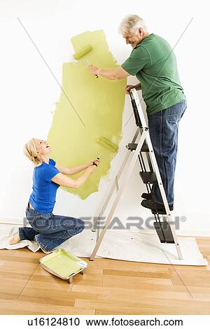 Stock Image - Middle-aged couple painting wall green with male on ladder.. Fotosearch