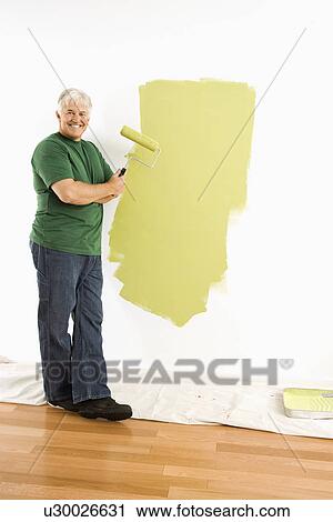 Stock Image - Middle-aged man painting wall green with paint roller smiling at viewer.. Fotosearch