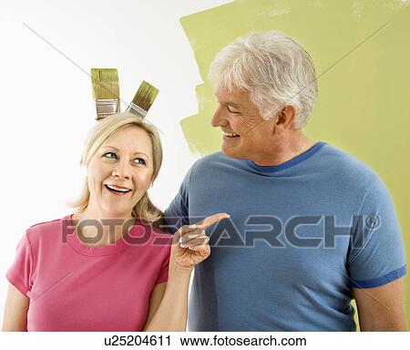 Portrait of happy adult couple standing in front of half-painted wall while man makes bunny ears with paintbrushes behind woman's head. View Large Photo Image Stock Image - Portrait of happy adult couple standing in front of half-painted wall while man makes bunny ears with paintbrushes behind woman's head.. Fotosearch
