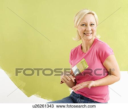 Stock Image - Portrait of smiling adult woman sitting in front of half-painted wall with paintbrush.. Fotosearch