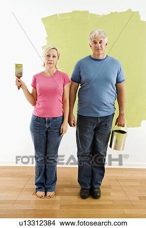 Portrait of unhappy adult couple standing in front of half-painted wall with paint supplies 'American Gothic' style View Large Photo Image Picture - Portrait of unhappy adult couple standing in front of half-painted wall with paint supplies 'American Gothic' style. Fotosearch