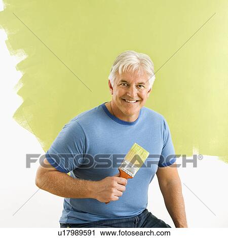 Stock Image - Smiling man sitting in front of half-painted wall with paintbrush. Fotosearch
