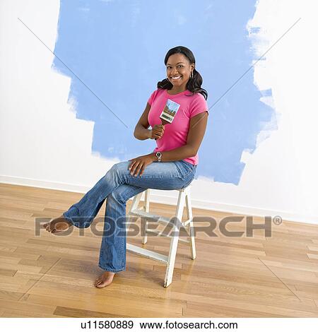 Stock Photo - Young woman sitting next to half-painted wall with paintbrush. Fotosearch