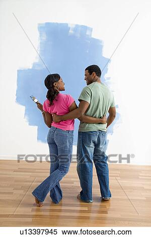Stock Photography - African American couple standing together next to half-painted wall discussing their work.. Fotosearch
