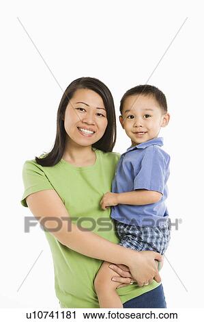 Stock Image - Asian mother holding son on hip smiling in front of white background.. Fotosearch