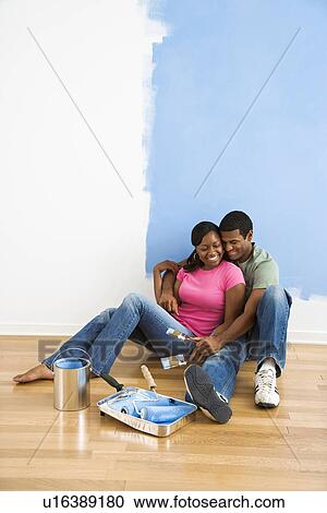 Stock Image - Couple sitting together relaxing next to half-painted wall and painting supplies. Fotosearch