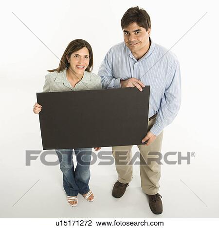 Stock Image - Couple standing smiling holding blank sign against white background. Fotosearch