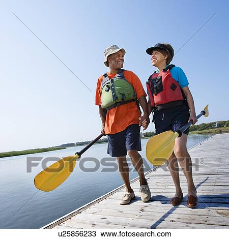 Stock Image - Couple walking dock carrying paddles. Fotosearch