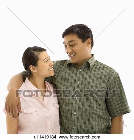 Picture - Mid adult Asian couple standing with arms around eachother and smiling at one another.. Fotosearch