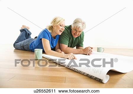Stock Image - Middle-aged couple lying on floor looking at and discussing architectural blueprints together.. Fotosearch