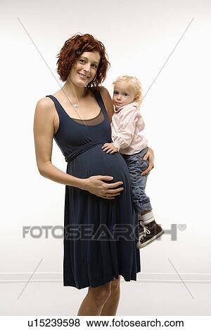 Stock Photo - Portrait of Caucasion mid-adult attractive pregnant woman standing, holding female toddler on hip and other hand on belly, looking at viewer and smiling.. Fotosearch