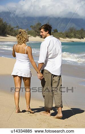 Stock Photography - Attractive couple holding hands and smiling at eachother walking on Maui, Hawaii beach.. Fotosearch