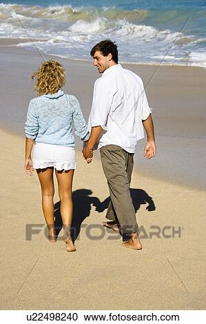 Stock Image - Attractive couple holding hands walking on beach smiling at eachother in Maui, Hawaii.. Fotosearch