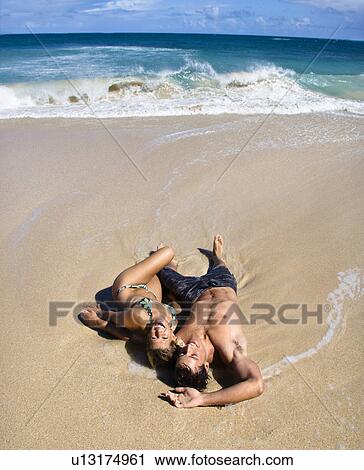 Stock Image - Attractive couple lying on beach smiling and laughing in Maui, Hawaii.. Fotosearch