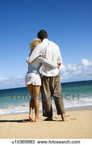 Stock Image - Attractive couple standing embracing looking out at ocean in Maui, Hawaii.. Fotosearch