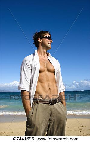 Stock Photograph - Portrait of attractive man standing with shirt unbuttoned wearing sunglasses on Maui, Hawaii beach.. Fotosearch