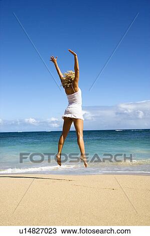 Stock Image - Pretty young woman jumping on Maui, Hawaii beach with arms raised into air.. Fotosearch
