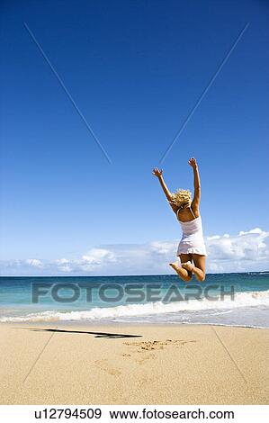 Stock Photo - Pretty young woman jumping on Maui, Hawaii beach with arms raised into air. Fotosearch