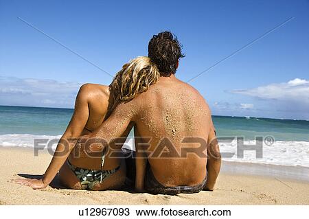 Stock Image - Rear view of couple sitting on Maui, Hawaii beach with woman leaning head on man's shoulder.. Fotosearch