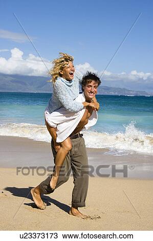 Stock Image - Woman riding piggyback on man while both smile and laugh on Maui, Hawaii beach. Fotosearch
