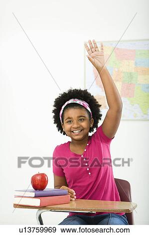 Stock Photo - African American girl sitting in school desk raising hand and smiling at viewer.. Fotosearch