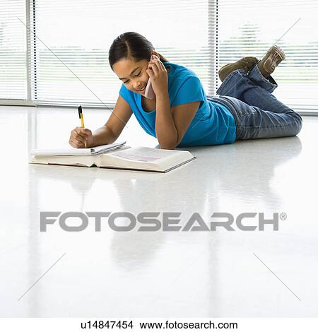 Girl lying on floor doing homework while talking on cell phone View Large Photo Image Picture - Girl lying on floor doing homework while talking on cell phone. Fotosearch