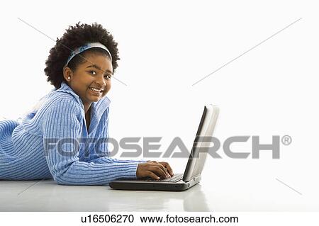 Stock Image - Side view of African American girl lying on floor using laptop and smiling at viewer.. Fotosearch