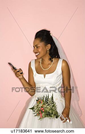 Stock Image - Mid-adult African-American bride looking at cellphone on pink background. . Fotosearch