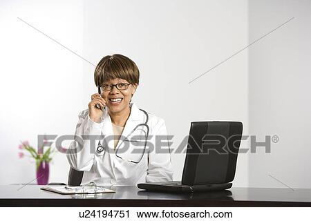 Stock Image - African American middle-aged female doctor sitting at desk working on laptop, talking on cell phone, smiling and looking at viewer.. Fotosearch