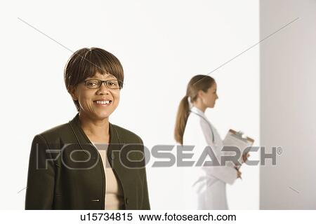 African American middle-aged female patient woman smiling looking at viewer with Caucasian mid-adult female doctor standing in background. View Large Photo Image Stock Photography - African American middle-aged female patient woman smiling looking at viewer with Caucasian mid-adult female doctor standing in background.. Fotosearch