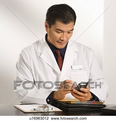 Stock Image - Asian American male doctor sitting at desk with charts using pda.. Fotosearch