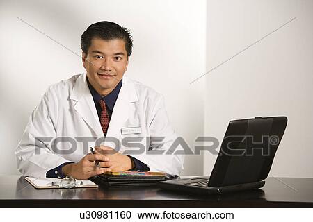Asian American male doctor sitting at desk with charts and laptop computer looking at viewer. View Large Photo Image Stock Image - Asian American male doctor sitting at desk with charts and laptop computer looking at viewer.. Fotosearch