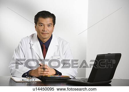 Stock Photography - Asian American male doctor sitting at desk with charts and laptop computer looking at viewer.. Fotosearch