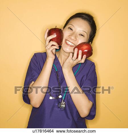Picture - Asian Chinese mid-adult female doctor holding two red apples up to face against yellow background smiling and looking at viewer.. Fotosearch