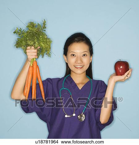 Picture - Asian Chinese mid-adult female doctor holding up bunch of carrots with one hand and red apple in other against blue background smiling and looking at viewer.. Fotosearch