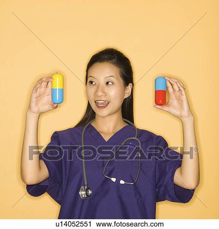 Stock Image - Asian Chinese mid-adult female doctor holding up giant pills against yellow background smiling.. Fotosearch