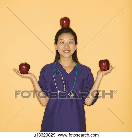 Stock Photo - Asian Chinese mid-adult female doctor holding two red apples in hands and balancing one on head against yellow background smiling and looking at viewer.. Fotosearch