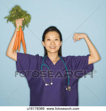 Stock Photograph - Asian Chinese mid-adult female doctor flexing muscle holding up bunch of carrots against blue background smiling and looking at viewer.. Fotosearch