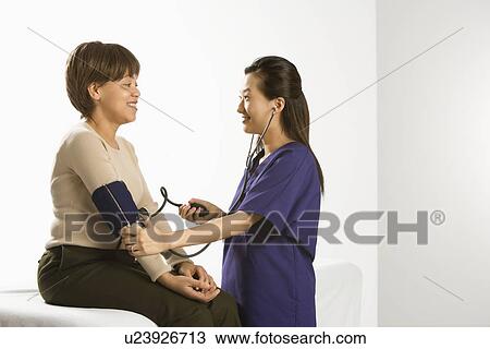 Stock Image - Asian Chinese mid-adult female medical practitioner checking blood pressure of African American middle-aged female patient.. Fotosearch