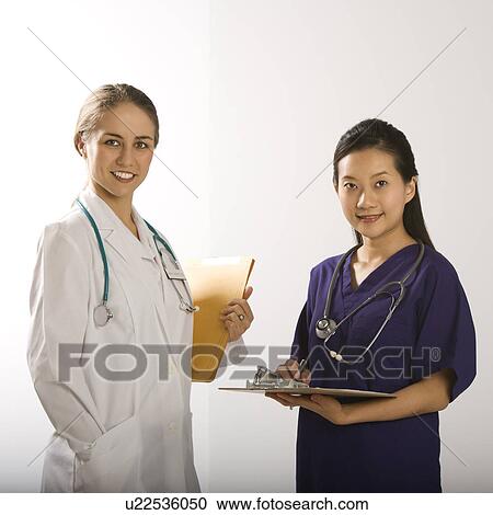 Stock Image - Caucasian mid-adult female doctor and Asian Chinese mid-adult female physician's assistant standing together smiling and looking at viewer.. Fotosearch