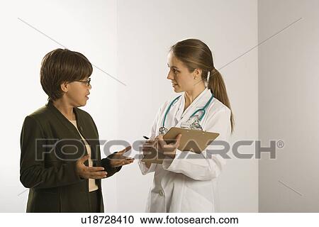 Stock Image - Caucasian mid-adult female doctor talking with African American middle-aged female patient.. Fotosearch
