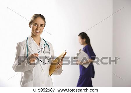 Caucasian mid-adult female doctor smiling and looking at viewer with Asian Chinese mid-adult female physician's assistant walking by in background. View Large Photo Image Stock Photo - Caucasian mid-adult female doctor smiling and looking at viewer with Asian Chinese mid-adult female physician's assistant walking by in background.. Fotosearch
